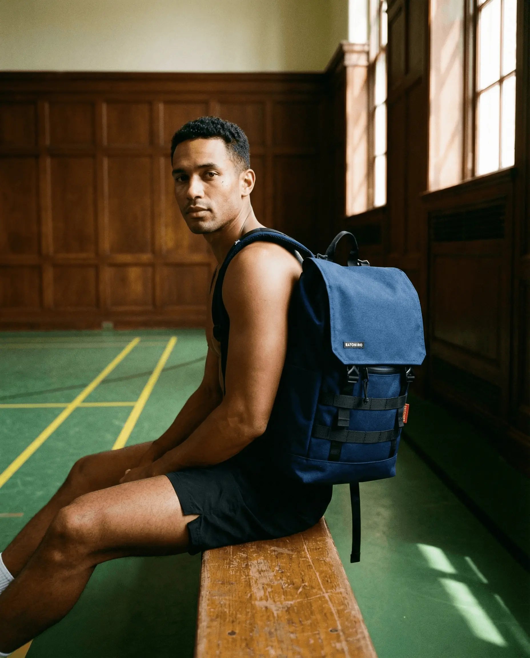Model sitting on a bench in an indoor court, wearing the Minato Lite 2.0 - Navy Blue backpack.