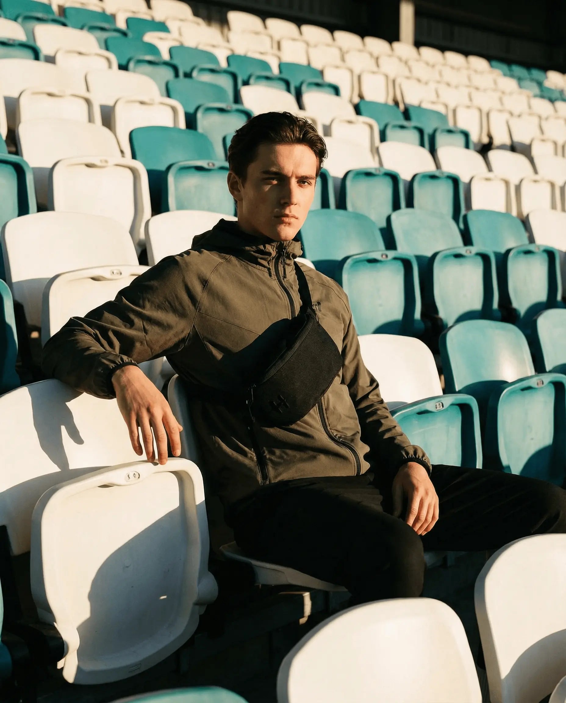 A model wearing the Sora 2.0 - Black Knight bag, sitting in a stadium with teal and white seats.