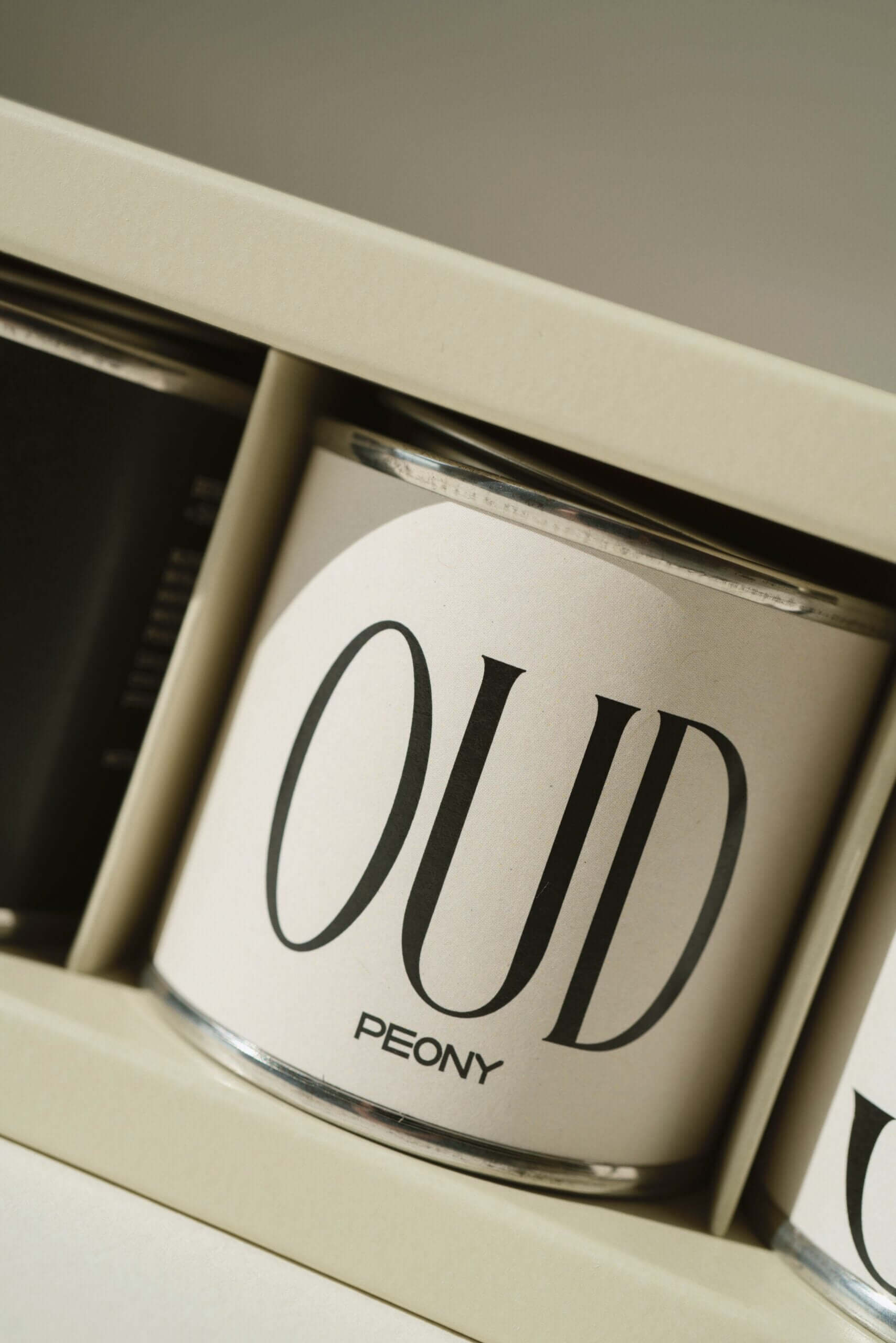 Close-up of mini metal tin candle labeled "OUD Peony" in elegant packaging, showcasing modern design.